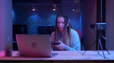 Kapalı neon arka plan ile dizüstü bilgisayarda video oyunları oynarken kulaklık dreadlocks ile genç çekici kadın blogger Closeup ateş