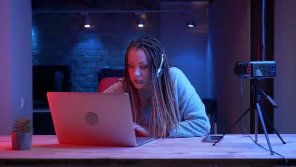 Primer plano de la joven atractiva blogger con rastas en los auriculares escribiendo en el portátil hablando en cámara con el fondo de neón en el interior — Vídeo de stock