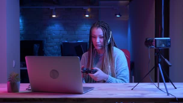 Tiro de close-up de jovem blogueiro feminino atraente com dreadlocks em fones de ouvido jogando jogos de vídeo no laptop com o fundo de néon dentro de casa — Vídeo de Stock