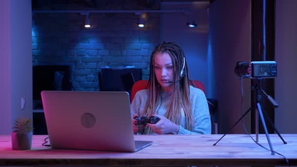 特写镜头的年轻有吸引力的女性博客与耳机的恐怖锁在笔记本电脑上玩视频游戏，并愉快地交谈，而流与霓虹灯背景在室内 — 图库视频影像