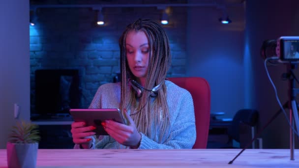 Gros plan shoot of young attractive female blogger with dreadlocks in headphones streaming live using the tablet and showing green chroma screen to the camera with the neon background inside — Video