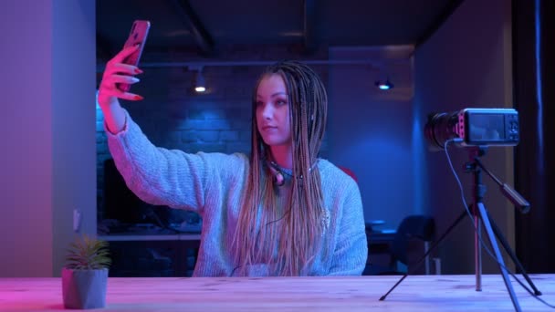 ドレッドロックを持つ若い魅力的な女性ブロガーのクローズアップ撮影は、屋内でネオンの背景とライブストリーミング携帯電話でセルフを取ります — ストック動画