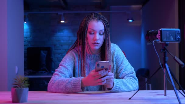 Съемки крупным планом молодой привлекательной блогерши с дредами на телефоне в прямом эфире с неоновым фоном в помещении — стоковое видео
