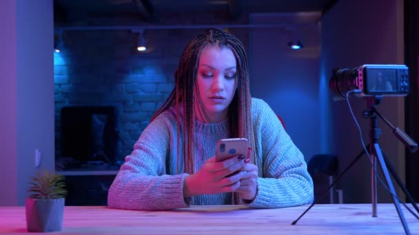 Gros plan shoot of young attractive female blogger with dreadlocks texting on the phone streaming live with the neon background à l'intérieur — Video