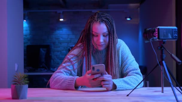 Sesión de primer plano de la joven bloguera atractiva con rastas mensajes de texto en el teléfono sonriendo alegremente en vivo con el fondo de neón en el interior — Vídeo de stock