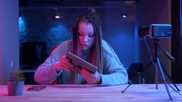 Sesión de primer plano de una joven bloguera atractiva con rastas jugando videojuegos en la tableta recibiendo transmisión emocional en vivo con el fondo de neón en el interior — Vídeo de stock