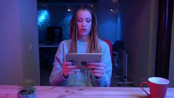 Sesión de primer plano de una joven bloguera atractiva con rastas jugando videojuegos en la tierra de la tableta mostrando la pantalla verde en vivo con el fondo de neón en el interior — Vídeos de Stock