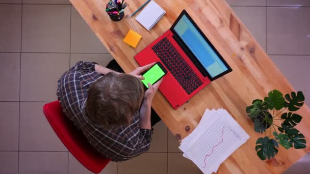Gros plan top shoot de jeune homme d'affaires caucasien tapant sur le téléphone assis devant l'ordinateur portable sur le bureau dans le bureau à l'intérieur — Video