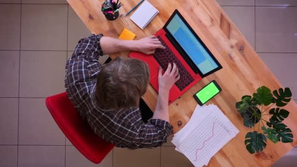 Närbild topp shoot av unga kaukasiska affärsman skriva på den bärbara datorn med en telefon på skrivbordet på kontoret inomhus — Stockvideo