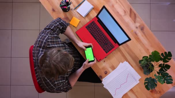 Primer plano sesión superior de joven hombre de negocios caucásico utilizando el teléfono con pantalla de croma verde sentado delante de la computadora portátil en el escritorio en la oficina en el interior — Vídeo de stock