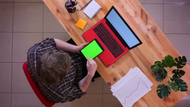 Close-up top shoot van jonge Kaukasische zakenman scrollen op de Tablet met groene Chroma scherm zittend voor de laptop in het kantoor binnenshuis — Stockvideo