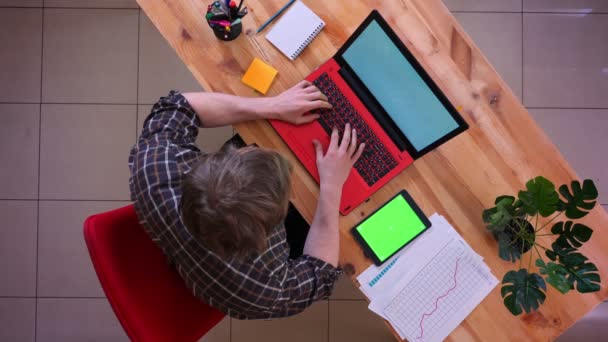 Primo piano ripresa superiore di giovane uomo d'affari caucasico digitando sul computer portatile con un tablet con schermo verde sulla scrivania in ufficio al chiuso — Video Stock