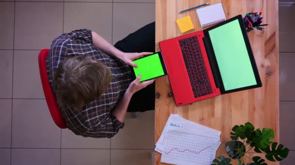 Κοντινά πλάνα του νεαρού καυκάσιου επιχειρηματία γραπτών μηνυμάτων στο tablet με πράσινη οθόνη αποχρώσεων μπροστά από το φορητό υπολογιστή στο γραφείο σε εσωτερικούς χώρους — Αρχείο Βίντεο