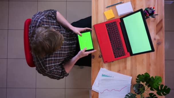 Hlavní natáčení mladého kavkazského podnikatele, který se nachází na tabletu se zelenou obrazovkou před laptopem v kanceláři v interiérech — Stock video