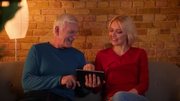 Primer plano de la sesión de ancianos feliz pareja usando la tableta sentada en el sofá interior en un acogedor apartamento — Vídeo de stock