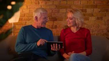 Tablet kullanarak üst düzey mutlu çift Closeup ateş ve rahat bir dairede kanepede otururken neşeyle gülümseyerek