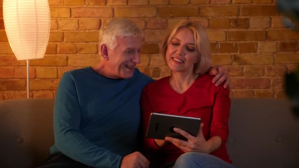 Primer plano de la sesión de ancianos feliz pareja navegar por la web en la tableta sonriendo alegremente sentado en el sofá interior en un acogedor apartamento — Vídeo de stock