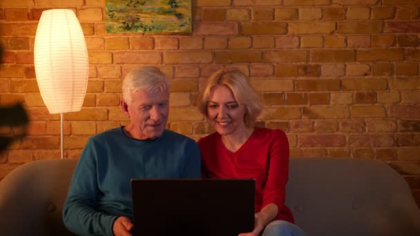 Primer plano de la sesión de ancianos feliz pareja usando el ordenador portátil sentado en el sofá interior en un acogedor apartamento — Vídeos de Stock