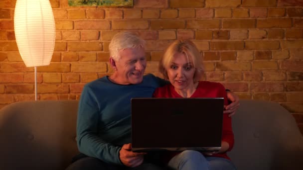 Tiro de close-up de casal feliz sênior usando as mídias sociais no laptop sorrindo alegremente sentado no sofá dentro de casa em um apartamento acolhedor — Vídeo de Stock