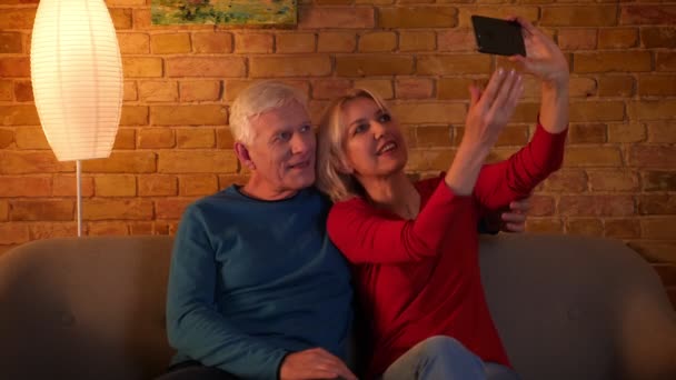 Съемки крупным планом пожилой счастливой пары, делающей селфи по телефону, сидящей на диване в уютной квартире — стоковое видео