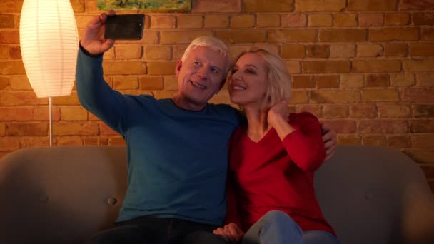 Close-up shoot van senior gelukkig paar het nemen van selfies op de telefoon glimlachend vrolijk zittend op de Bank binnenshuis in een gezellig appartement — Stockvideo