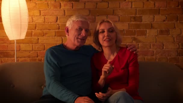 Närbild shoot of Senior lyckligt par tittar på en TV-film leende glatt sitter på soffan inomhus i en mysig lägenhet — Stockvideo