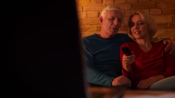 Съемки крупным планом счастливой пожилой пары, смотрящей документальный фильм, весело улыбающейся сидя на диване в уютной квартире — стоковое видео