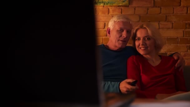 Primer plano de la sesión de ancianos feliz pareja viendo un documental de televisión sonriendo alegremente sentado en el sofá interior en un acogedor apartamento — Vídeos de Stock