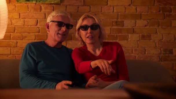 Plan rapproché d'un couple heureux âgé regardant un film 3D à la télévision avec joie assis sur le canapé à l'intérieur dans un appartement confortable — Video