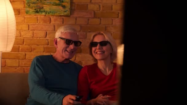 Primer plano de la sesión de ancianos feliz pareja viendo una película en 3D en la televisión riendo alegremente sentado en el sofá dentro de un acogedor apartamento — Vídeos de Stock