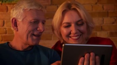 Tablette görüntülü görüşme olan yaşlı mutlu çiftin yakın çekim çekimi, rahat bir dairede kanepede neşeyle otururken gülümsüyor
