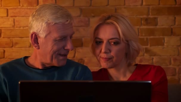 Primer plano sesión de ancianos feliz pareja viendo una película en el ordenador portátil sentado en el sofá en el interior en un acogedor apartamento — Vídeo de stock