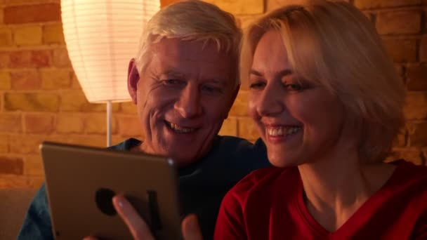 Close-up shoot van leeftijd gelukkig paar met een video-oproep op de Tablet en communiceren vrolijk zittend op de Bank binnenshuis in een gezellig appartement — Stockvideo