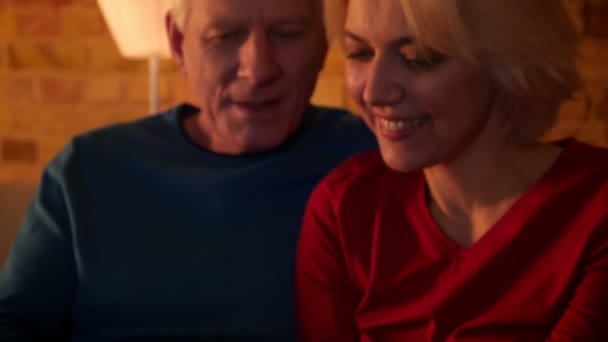 Tiro de close-up de casal feliz envelhecido usando o tablet sentado no sofá dentro de casa em um apartamento acolhedor — Vídeo de Stock