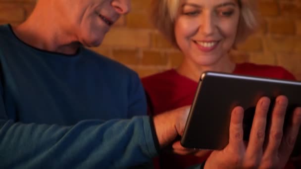 Close-up shoot van leeftijd gelukkig paar met behulp van de Tablet zittend op de Bank rust samen binnen in een gezellig appartement — Stockvideo