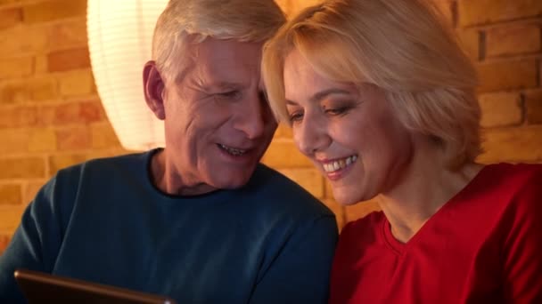 Gros plan shoot de vieux couple heureux regardant une comédie sur la tablette riant joyeusement assis sur le canapé à l'intérieur dans un appartement confortable — Video