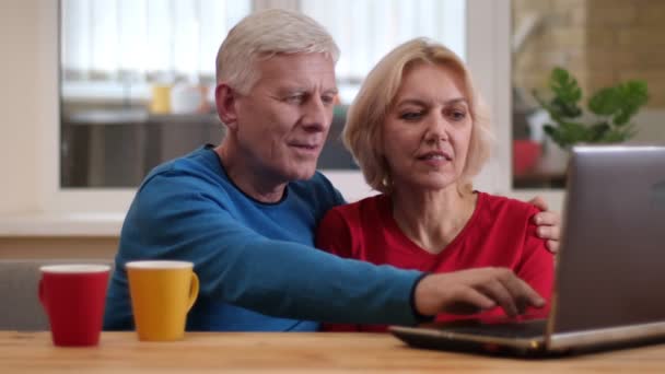 Primer plano de la sesión de ancianos feliz pareja utilizando el ordenador portátil en el escritorio con tazas de té en el interior en un acogedor apartamento — Vídeo de stock