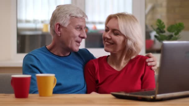 Primer plano de la sesión de ancianos feliz pareja utilizando el ordenador portátil en el escritorio con tazas de té en el interior en un acogedor apartamento en un día — Vídeos de Stock