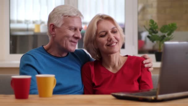 Zbliżenie strzelać starszy szczęśliwy para za pomocą laptopa na biurku z filiżanek z herbaty uśmiechnięty szczęśliwie w pomieszczeniu w przytulnym mieszkaniu — Wideo stockowe