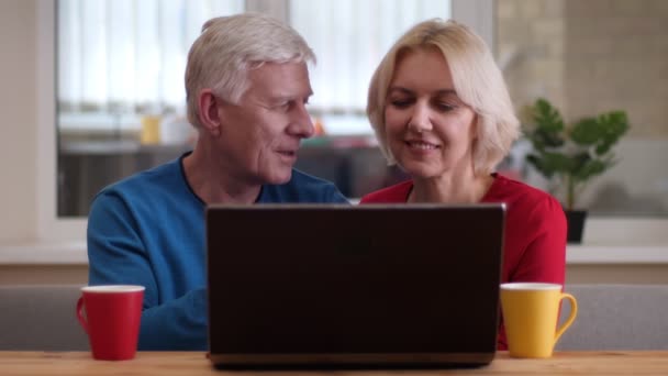 Sesión de primer plano de la pareja feliz de edad utilizando el ordenador portátil en el escritorio con tazas de té en el interior en un acogedor apartamento — Vídeo de stock