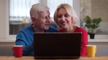 Rahat bir dairede kapalı masada çay ile bardak ile dizüstü bilgisayar kullanarak yaşlı mutlu çift Closeup ateş