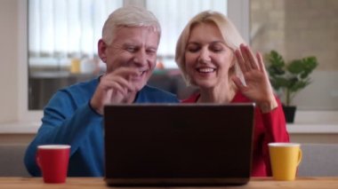 Masanın üzerinde çay ile bardak ile dizüstü bilgisayarda bir video arama sahip yaşlı mutlu çift Closeup ateş ve rahat bir dairede merhaba kapalı sallayarak