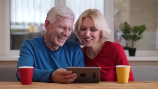 Tiro de close-up de casal feliz envelhecido ter uma chamada de vídeo do tablet com copos com chá na mesa dentro de casa em um apartamento acolhedor — Vídeo de Stock