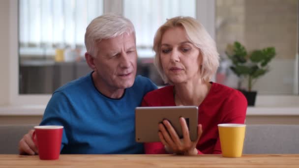 Sesión de primer plano de la pareja feliz anciana navegando en la tableta con tazas con té en el escritorio en un acogedor apartamento — Vídeos de Stock