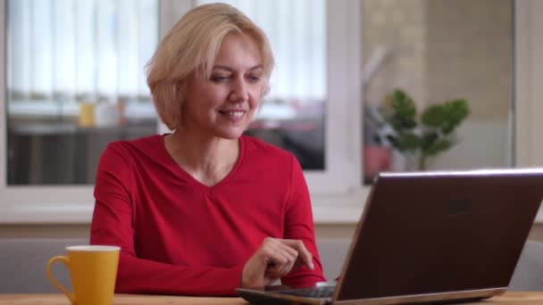 Tiro de close-up de mulher caucasiana envelhecida digitando no laptop dentro de casa em um apartamento acolhedor — Vídeo de Stock