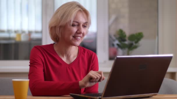 Крупним планом знімок літньої білої жінки, що має відеодзвінок на ноутбук, посміхаючись і розмовляючи щасливо в приміщенні в затишній квартирі — стокове відео