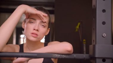 Genç çekici uygun kadın yürüyüş closeup ateş ve kapalı spor salonunda yorgunluk ile kamera düz bakarak ağırlık kaldırmaya hazırlanıyor