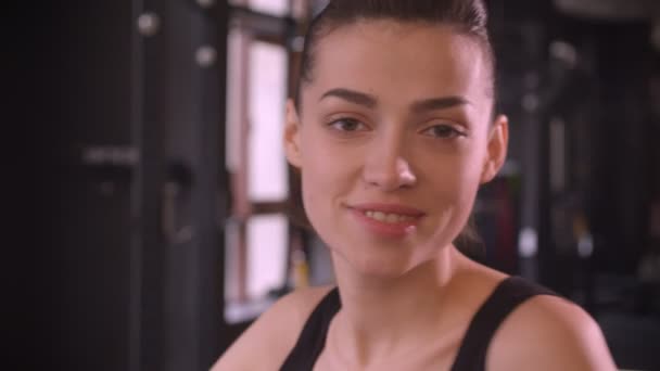 Primer plano de la joven atleta atractiva hembra con cola de caballo haciendo ejercicio en el orbitrek sonriendo felizmente y mirando a la cámara en el gimnasio en el interior — Vídeos de Stock