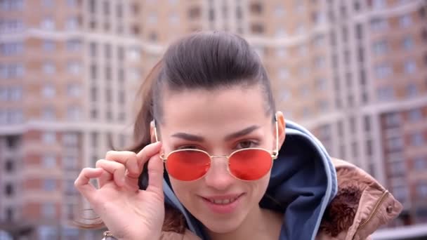 Primer plano de la joven atractiva chica caucásica en gafas de sol rojas con cola de caballo sonriendo y mirando a la cámara en la ciudad urbana al aire libre — Vídeo de stock