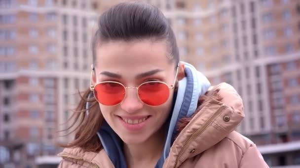 Close-up tiro de jovem atraente menina caucasiana na moda em óculos de sol vermelhos com rabo de cavalo sorrindo e olhando para a câmera de pé no casaco na cidade urbana ao ar livre — Vídeo de Stock
